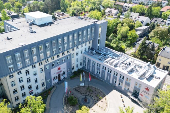 Widok z lotu ptaka na kompleks budynków Bonifraterskiego Centrum Medycznego. Wielokondygnacyjny obiekt ma biało-szarą elewację i liczne okna. Przed głównym wejściem znajduje się półkolisty podjazd, niewielki teren zielony z pomnikiem oraz maszty z flagami. Na dachach widoczne są instalacje techniczne i wentylacyjne. Budynek otaczają drzewa, zabudowa mieszkaniowa i ulice.